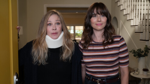 Two women standing in the entryway of a house; one wears a neck brace and dark sweater, the other wears a striped shirt. They stand near an open yellow door, with a staircase and potted plant visible in the background.