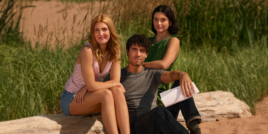 Three young adults sitting on rocks outdoors with tall green grass and sand in the background, enjoying a sunny day in a natural setting.