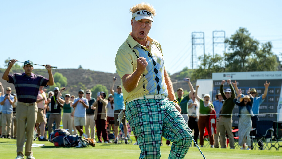 Golfer celebrating on green with fist pump, wearing plaid pants and visor, as cheering crowd watches in a sunny outdoor golf course setting with scoreboard and hills in background.