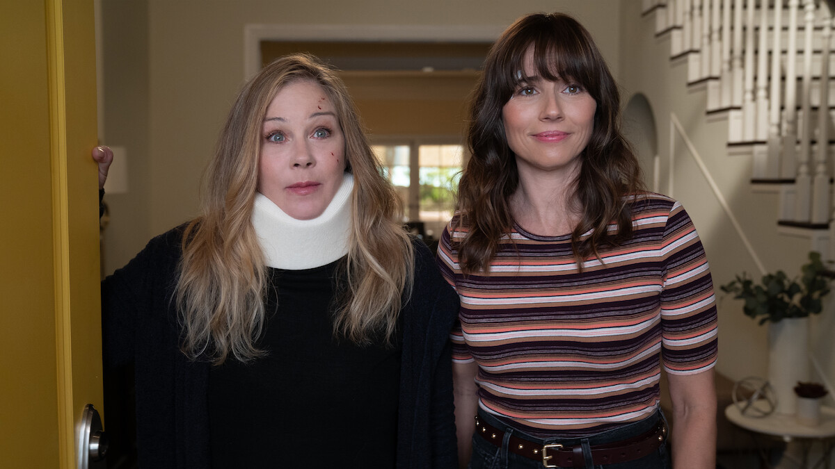 Two women standing in the entryway of a house; one wears a neck brace and dark sweater, the other wears a striped shirt. They stand near an open yellow door, with a staircase and potted plant visible in the background.