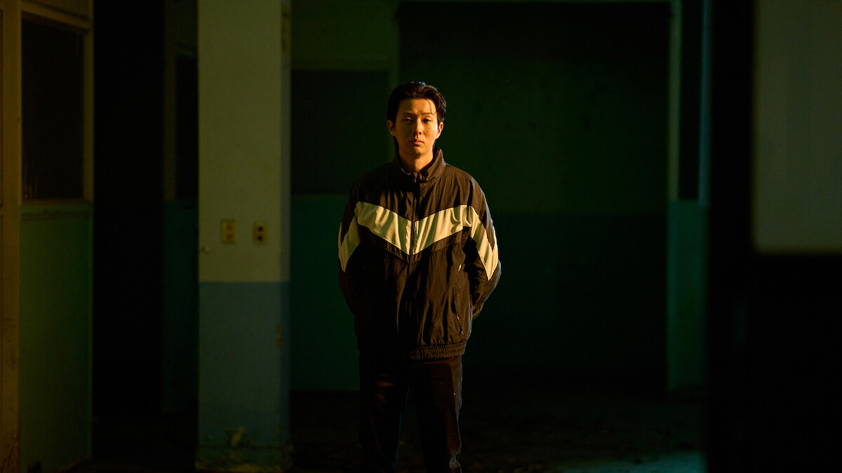  Choi Woo-shik in the series A Killer Paradox stands in a track jacket in a darkened corridor with evening light hitting the right side of his face.