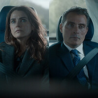 Two people, a woman and a man, sit side by side in a car wearing seatbelts. Both appear serious and are dressed in business attire, with a blurred background visible through the car windows.