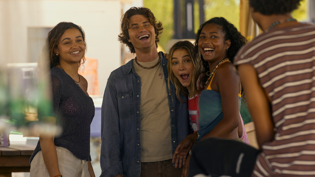 Four young adults stand closely together, smiling and laughing outdoors in a casual, sunny setting, with another person partially visible in the foreground. The group appears to be enjoying a relaxed, friendly moment together.