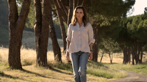 A woman walks alone on a dirt path through a sunlit forest clearing, surrounded by tall trees and grass on a clear day.