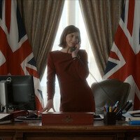 Woman in a formal red dress stands at an office desk, speaking on a corded phone. The room features large windows, two British flags, and office supplies, suggesting a government or diplomatic setting.