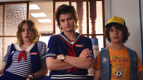  Three teenagers stand indoors; two wear matching blue sailor-style uniforms, appearing to work at an ice cream shop, while the third wears a colorful t-shirt and cap. Their expressions are serious, creating a tense mood.