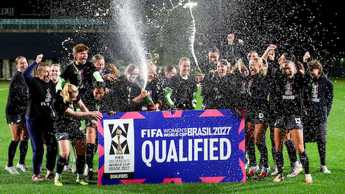 Women's soccer team celebrates at night on a field, spraying champagne and cheering behind a large sign reading "FIFA Women's World Cup Brasil 2027 Qualified." Bright stadium lights and festive atmosphere.