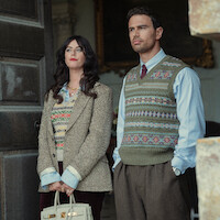 A woman and man stand in a doorway wearing vintage-style clothing with patterned sweaters; the setting appears formal and indoors, with a sophisticated, historic atmosphere.