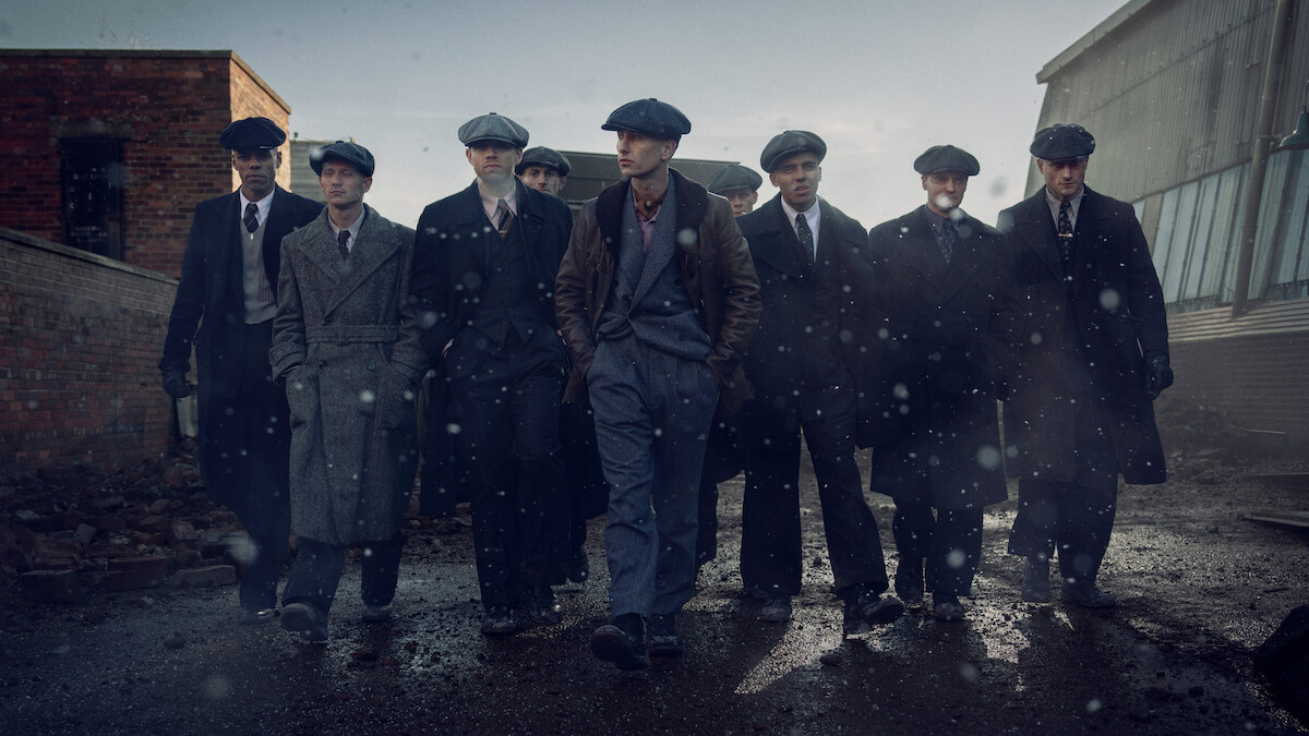 Eight men in vintage suits and flat caps walk together on a wet, gritty street in an industrial urban area, with old brick and metal buildings in the background under a gray, overcast sky.