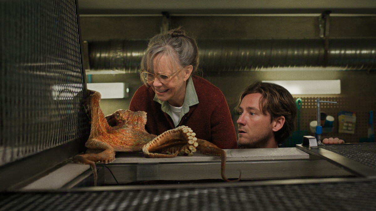 An older woman and a younger man observe an octopus on a metal surface in a laboratory or aquarium setting, with industrial pipes and equipment in the background.