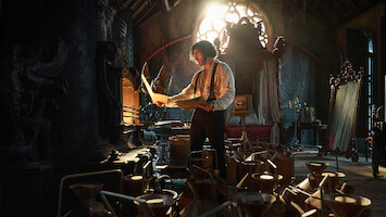 A man studies papers in a dark, ornate room filled with scientific equipment and warm, dramatic light streaming through a stained-glass window, creating a mysterious and intense mood.