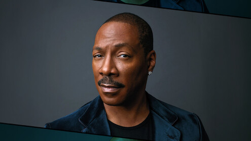 Eddie Murphy with short hair, mustache, wearing a dark velvet jacket and diamond earring, poses against a plain dark background, with a serious and confident expression. Studio lighting highlights his face.