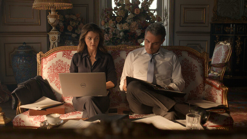 Two people sit on an ornate sofa in an elegant, traditional room with paperwork, a laptop, flowers, and decorative objects, appearing to work seriously together.