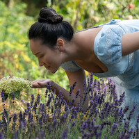 Woman in a blue dress bending down to smell flowers in a lush garden filled with lavender and greenery on a sunny day.