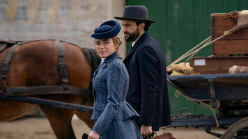 A woman and man in period clothing stand by a horse-drawn wagon with luggage, outdoors in a historical setting, suggesting a travel or arrival scene.