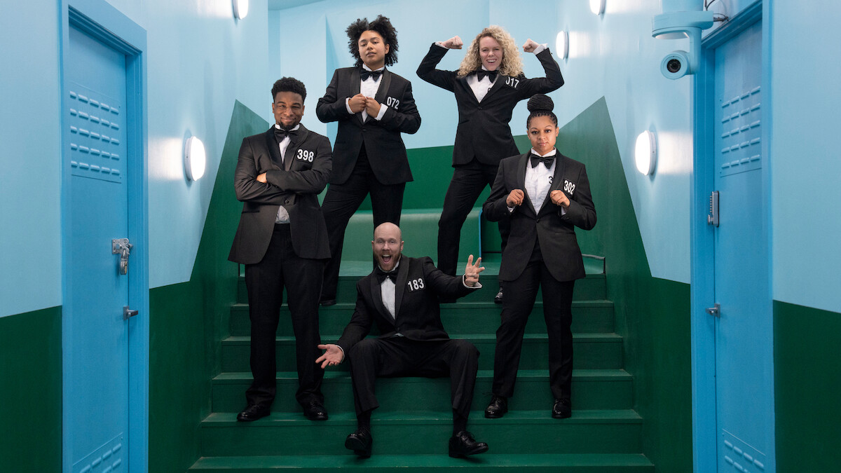Five people in black suits pose confidently on green stairs in a brightly lit, blue-walled hallway, creating a playful, strong, and upbeat atmosphere.