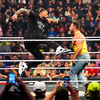 Two wrestlers in a crowded arena; one jumps with a knee strike toward the other inside a wrestling ring, surrounded by ropes, with fans watching and capturing the action on their phones in the background.