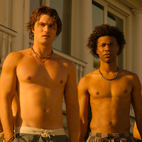 Two young men standing shirtless and wet outdoors, wearing swim trunks and necklaces, in front of a white building with columns and large windows, illuminated by golden sunlight.