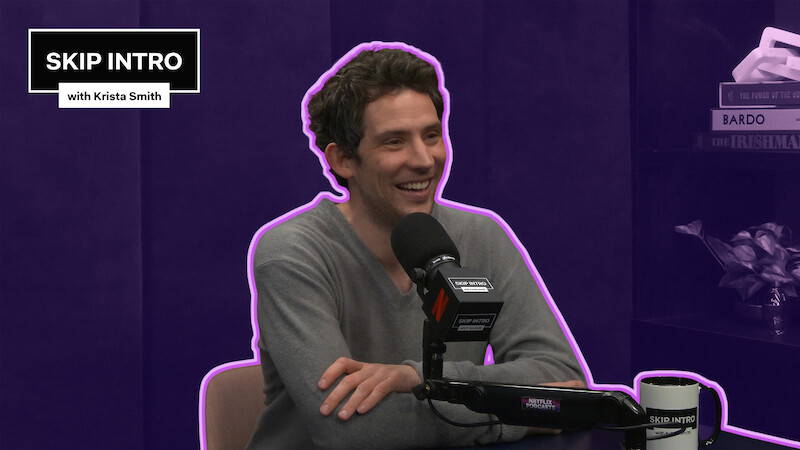Josh O'Connor smiles while speaking into a podcast microphone in a purple studio, outlined with a neon accent, with a “Skip Intro with Krista Smith” graphic in the corner.