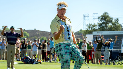 Golfer celebrating on green with fist pump, wearing plaid pants and visor, as cheering crowd watches in a sunny outdoor golf course setting with scoreboard and hills in background.