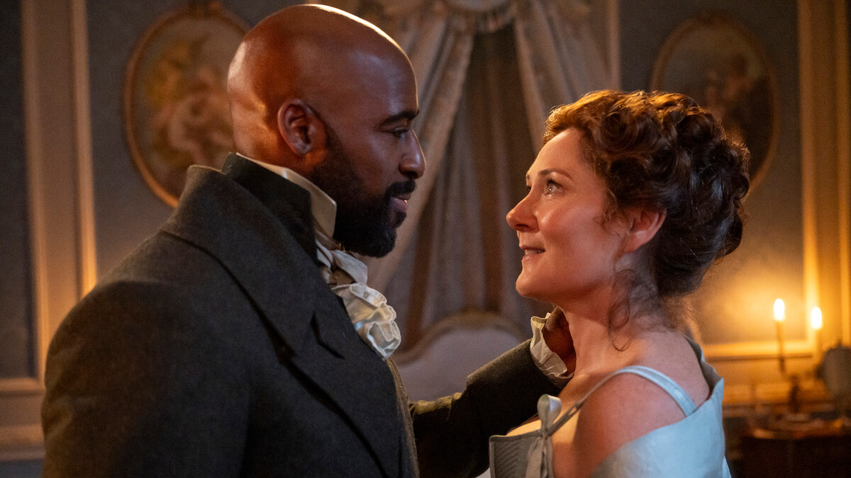 A man and woman in historical clothing gaze at each other in an elegantly decorated, candlelit room with ornate walls and portraits, suggesting a romantic or emotional moment in a period drama setting.