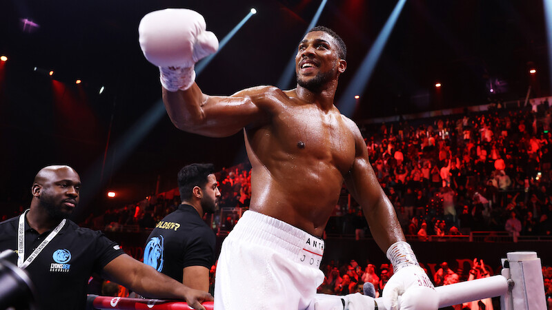 A victorious boxer with white gloves and shorts smiles in a brightly lit boxing ring, surrounded by a cheering crowd and security staff, creating an energetic and triumphant atmosphere.
