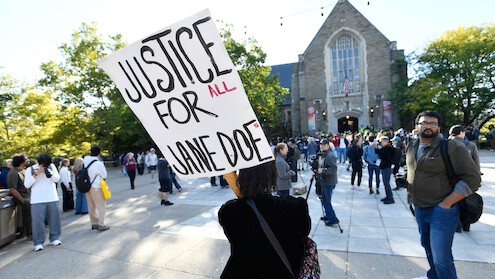 Person holds "Justice for Jane Doe" sign at outdoor protest in front of a large stone building, with a crowd of people gathered around under a sunny sky with trees in the background.