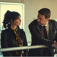 A woman in a leather jacket and scarf stands next to a man in a suit and tie. They are indoors, leaning on a metal railing, looking at each other in a serious conversation in a modern, softly lit room.