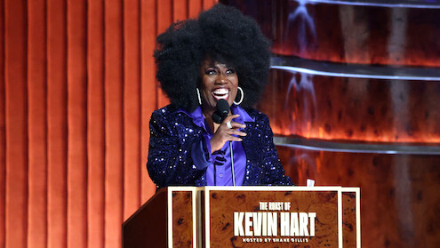 Sheryl Underwood with a large afro hairstyle and sparkly purple jacket speaks at a podium labeled "The Roast of Kevin Hart," on a stage with wood paneling and warm lighting.