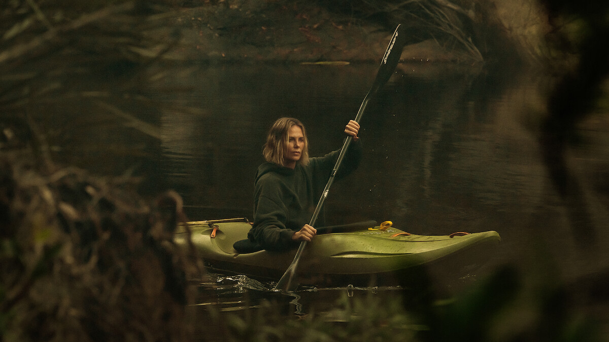 A person paddling a green kayak on a calm, forested river surrounded by trees and branches, wearing a dark hoodie and looking ahead with a serious expression, in a natural outdoor setting.