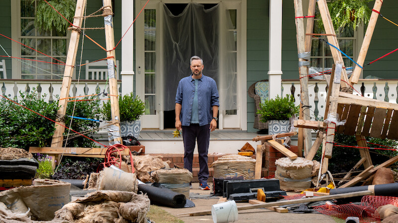 A man stands on a porch surrounded by construction materials, debris, and wooden scaffolding in front of a house. The setting suggests renovation work in progress, with scattered tools and building supplies.