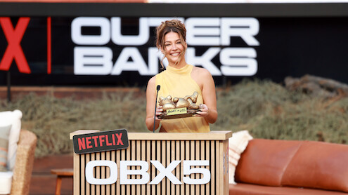 Smiling woman in yellow dress holding award, standing at a Netflix OBX5 podium outdoors with Outer Banks signage and casual seating in the background.
