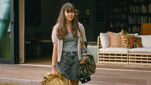 Young woman with long hair stands outside a cozy, modern house holding a coat, bag, and camera. The relaxed setting features a wicker sofa and bookshelves, evoking a calm, casual, and welcoming atmosphere.