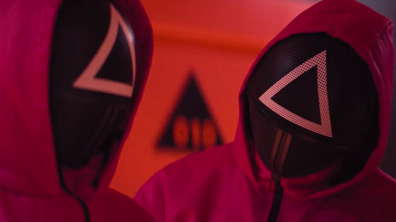 Duas figuras mascaradas com capacetes de esgrima se olham em um ambiente com iluminação vermelha