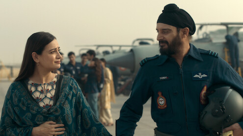 A woman and a male Air Force officer in uniform walk together on an airbase, with military aircraft and other personnel visible in the background, evoking a sense of camaraderie and support.