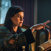 Woman in a dark robe sits on an ornate chair indoors, raising her hand as if casting a spell, with warm, moody lighting and a partially visible map in her lap.