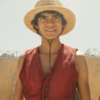 Young person smiling, wearing a straw hat and red vest, standing outdoors on a sunny day with blurred buildings in the background.