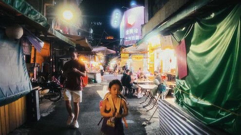 Child and adult walk through a bustling, neon-lit night market with food stalls, tables, and people dining; the scene is lively and colorful, evoking a vibrant urban atmosphere.