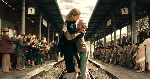 Barbara Ronchi and Christian Cervone hug in a train station on the train trains surrounded by people clapping in an image from the 1940s period film 'The Children's Train.'