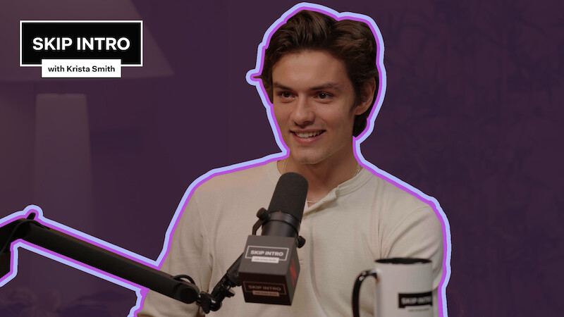 Louis Partridge smiles while speaking into a microphone during an interview on "Skip Intro with Krista Smith."