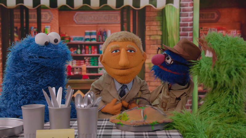 Four colorful puppets sitting at a checkered table in a cheerful, cozy café setting, with kitchen utensils and a pie.