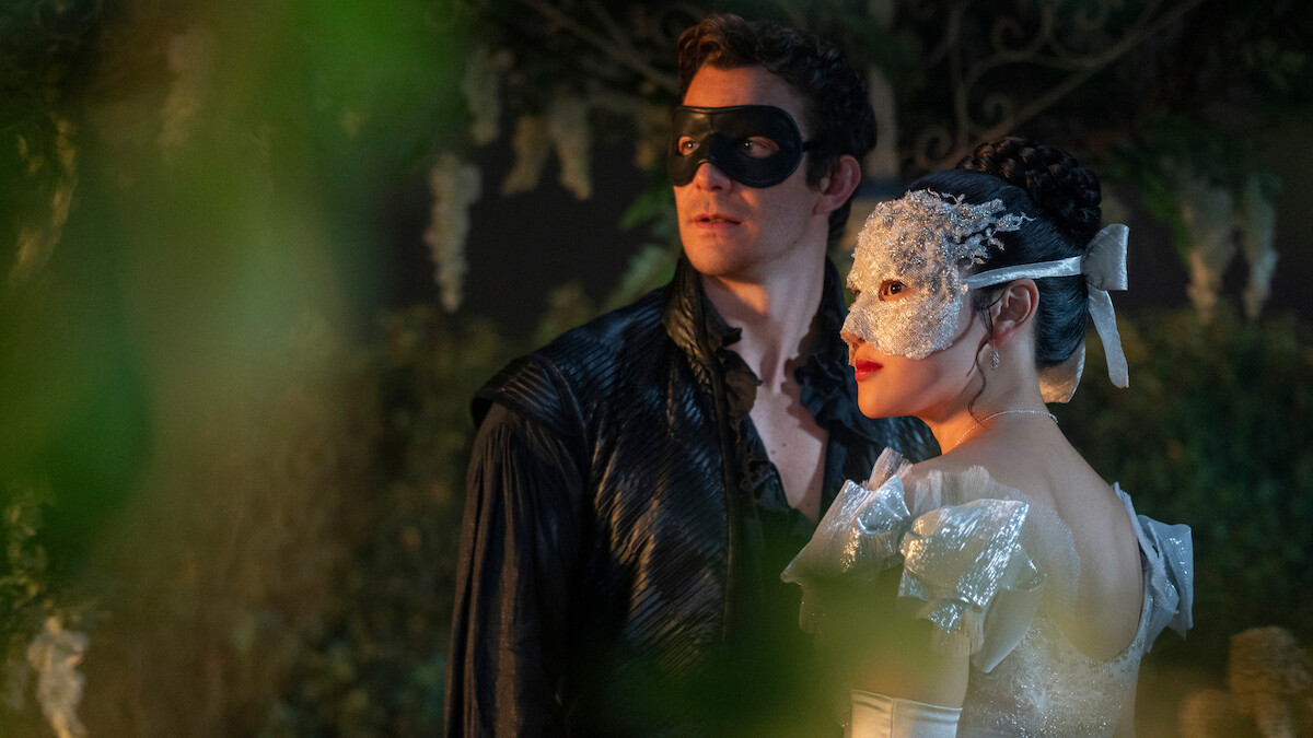 A man and woman in elaborate masquerade masks and formal costumes stand close together in a lush, evening garden setting with greenery and soft lighting.