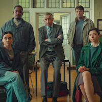Five people, three seated and two standing, pose indoors in a well-lit room with wooden floors and stained-glass windows, dressed in formal and casual clothing, appearing serious and focused.