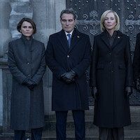 Three people in dark formal coats and gloves stand outside in front of an old stone building and ornate doors, posing seriously for a photograph in a somber or official setting.