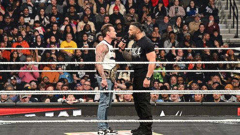 Two wrestlers face off in a packed arena wrestling ring, one holding a championship belt and the other speaking into a microphone as a large audience watches the dramatic confrontation.