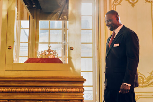 Um homem negro, alto e de cabelos raspados usa terno preto e observa uma coroa dentro de seu recipiente, em um museu