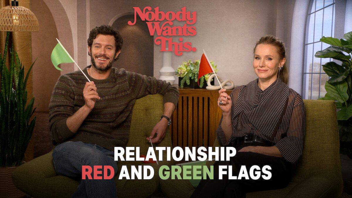 Adam Brody (Noah) and Kristen Bell (Joanne) sit on a couch in a cozy living room, smiling and holding green and red flags, with text about "Relationship Red and Green Flags," creating a lighthearted and conversational mood.