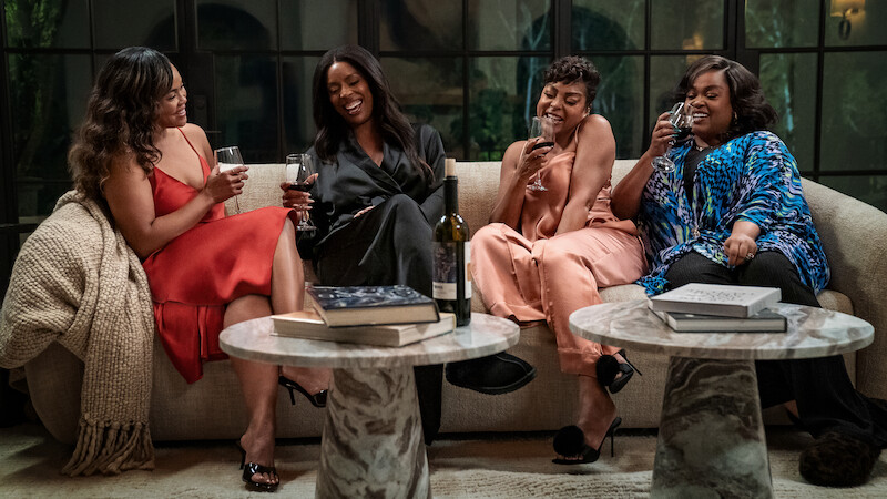 Four women sit on a beige couch in an elegant living room, laughing and enjoying glasses of wine, with books, throw blanket, and a wine bottle on marble coffee tables, creating a cozy, relaxed, and joyful atmosphere.