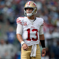 Football player wearing a San Francisco 49ers uniform and helmet stands on the field during a game, looking to the side, with a blurred stadium crowd in the background.