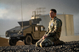 A soldier in camouflage uniform kneels on rocky ground near a military vehicle, with an industrial tank in the background under a cloudy sky, capturing a contemplative and solemn mood.